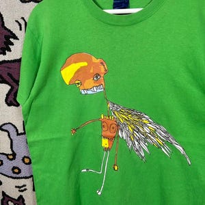 May include: A green t-shirt featuring a whimsical illustration of a creature with a brown head, yellow and white wings, and a long, thin body. The artwork has a cartoonish style with a playful and unique design.