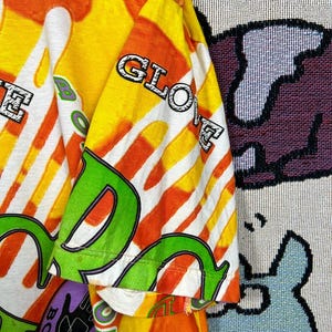 May include: A vibrant, short-sleeved shirt with a bold, colorful design. The shirt features the text "BODY GLOVE" in a mix of green, white, and purple, set against a backdrop of orange, yellow, and white stripes. The sleeve displays the word "GLOVE".