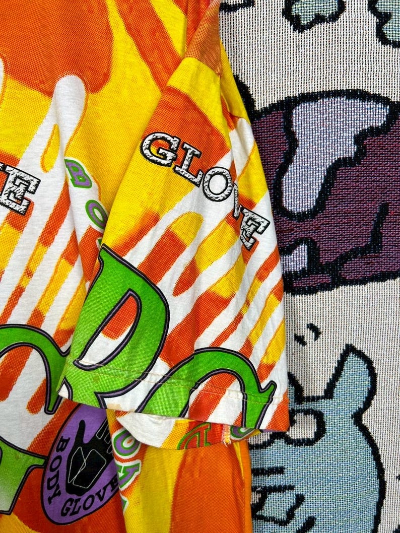 May include: A vibrant, short-sleeved shirt with a bold, colorful design. The shirt features the text "BODY GLOVE" in a mix of green, white, and purple, set against a backdrop of orange, yellow, and white stripes. The sleeve displays the word "GLOVE".