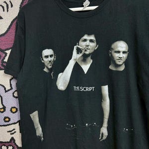 Puede incluir: Camiseta negra con una imagen en blanco y negro de la banda The Script. Los miembros de la banda están en una foto de grupo, con el nombre de la banda impreso en blanco. La camiseta tiene cuello redondo y mangas cortas.