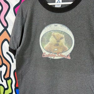 May include: A charcoal grey ringer t-shirt with black trim on the collar and sleeves. The center of the shirt features a circular graphic of a gopher inside a space helmet with the text Caddyshack printed in red lettering below the image.