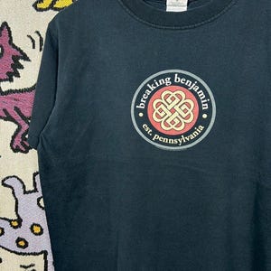 May include: Black t-shirt featuring a circular logo with the text "breaking benjamin" and "est. pennsylvania". The logo has a red and gold design. The shirt is hanging on a white hanger.