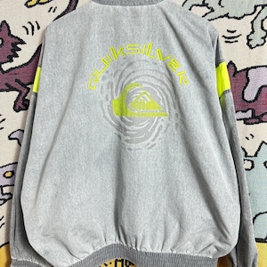 Può includere: Giacca grigia Quiksilver con collo alto e chiusura a zip. La parte posteriore presenta un logo e testo verde neon. Strisce verde neon accentuano le maniche. La giacca ha orlo e polsini a coste.