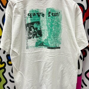 May include: White t-shirt with a green graphic print that says "Have fun!" and features a photo of a person with a microphone. The text "With Hansons" is printed below the graphic.
