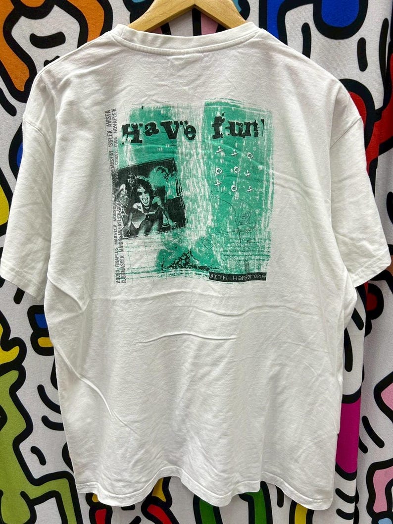 May include: White t-shirt with a green graphic print that says "Have fun!" and features a photo of a person with a microphone. The text "With Hansons" is printed below the graphic.