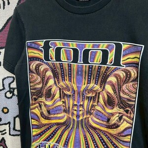 Może przedstawiać: Czarny t-shirt z kolorowym wzorem graficznym. Słowo "TOOL" jest wyświetlane białymi literami blokowymi. Grafika przedstawia psychodeliczny obraz z tekstem "10,000 days" na dole.