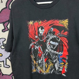 Puede incluir: Camiseta negra con una ilustración de cómic de Spawn, un superhéroe con capa roja y cadenas. El gráfico es detallado con colores vibrantes y poses dinámicas, típico del arte del cómic.
