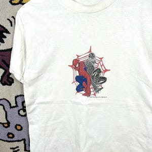 May include: White t-shirt with a graphic of Spider-Man in red, blue, and black, set against a spiderweb. The text "SPIDER-MAN" is printed below. The shirt has a crew neck and short sleeves.