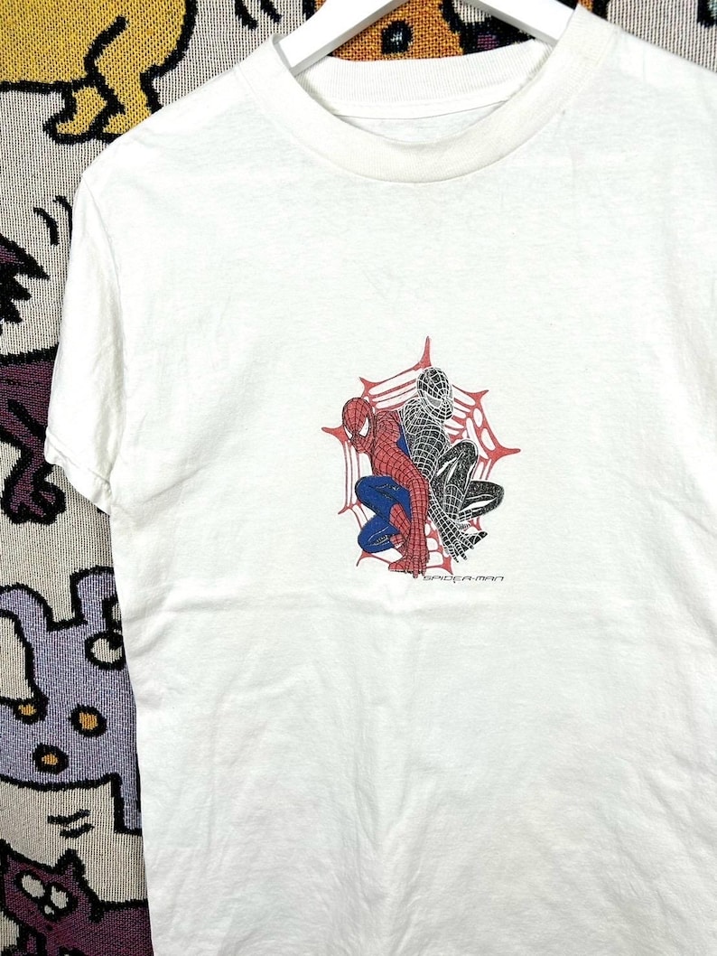 May include: White t-shirt with a graphic of Spider-Man in red, blue, and black, set against a spiderweb. The text "SPIDER-MAN" is printed below. The shirt has a crew neck and short sleeves.