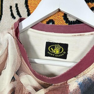 May include: A vintage t-shirt with a unique abstract pattern in shades of brown, beige, and blue. The shirt has a burgundy neckline and the "Body Glove" logo on a black tag. The shirt is hanging on a white hanger.