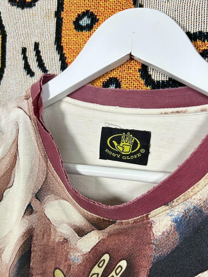May include: A vintage t-shirt with a unique abstract pattern in shades of brown, beige, and blue. The shirt has a burgundy neckline and the "Body Glove" logo on a black tag. The shirt is hanging on a white hanger.