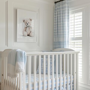May include: A white cot with blue and white checked bedding and a matching blanket. A framed watercolour print of a teddy bear hangs above the cot. Blue and white plaid curtains and a window with shutters are visible.
