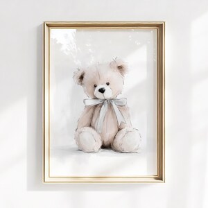 May include: A framed watercolour print of a teddy bear. The bear is light brown with a grey bow around its neck. The artwork is in a gold frame and is displayed against a white wall.