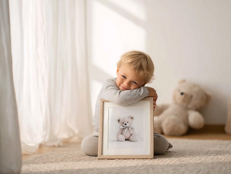 May include: A framed print of a teddy bear with a bow. The print is held by a child. The print has a light wooden frame. A stuffed teddy bear is in the background. The room has a neutral colour palette.