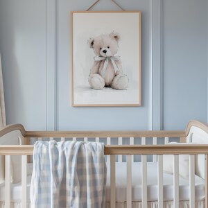May include: A framed watercolour print of a teddy bear with a pale blue ribbon, hanging above a cot with a blue and white checked blanket. The cot is made of light wood and has white bedding.