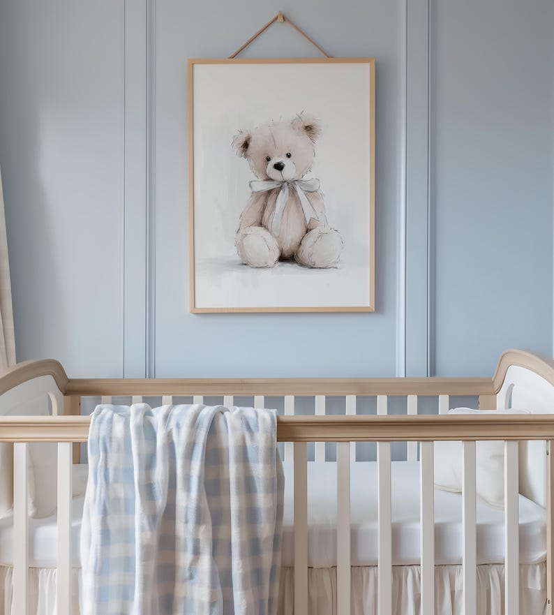 May include: A framed watercolour print of a teddy bear with a pale blue ribbon, hanging above a cot with a blue and white checked blanket. The cot is made of light wood and has white bedding.
