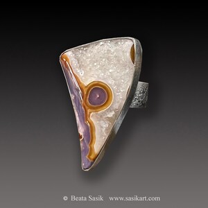 May include: A silver ring featuring a triangular shaped stone set in the centre. The stone is a mix of white, brown, and purple colours with a circular pattern.