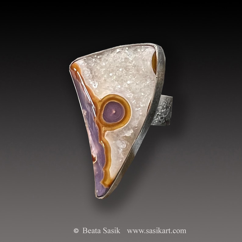 May include: A silver ring featuring a triangular shaped stone set in the centre. The stone is a mix of white, brown, and purple colours with a circular pattern.