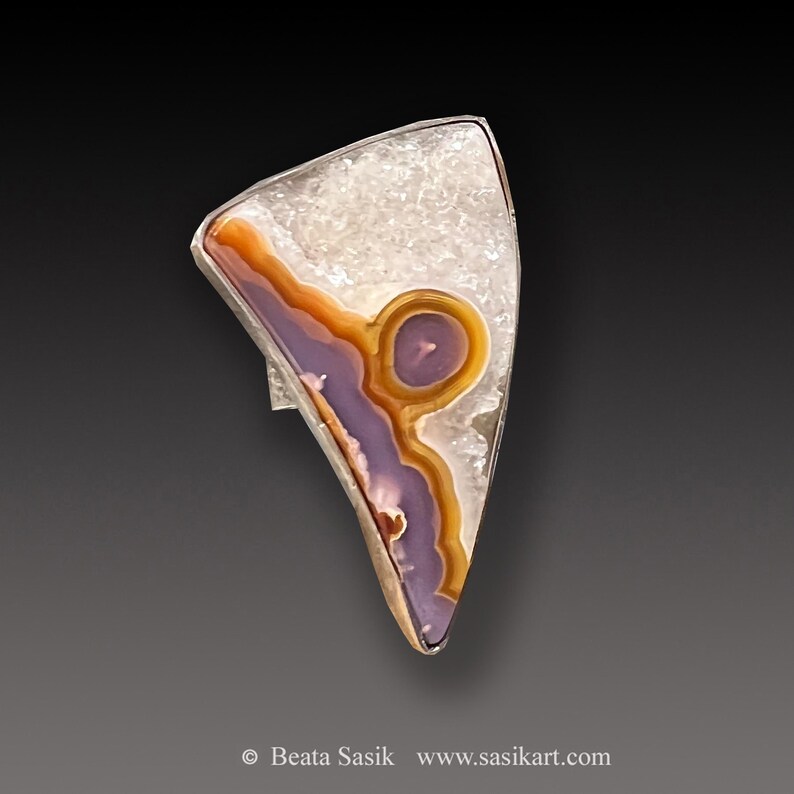 May include: A silver ring featuring a purple, orange and white agate stone. The stone is set in a triangular shape and displays a distinctive pattern.