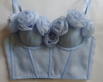 May include: A light blue corset top with a sweetheart neckline and a bustier style. The top is adorned with large, light blue fabric flowers.