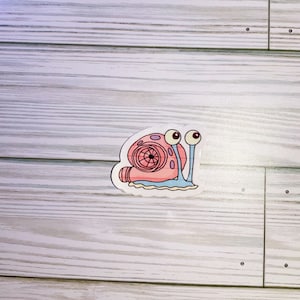 May include: A sticker featuring a cartoon snail with a turbocharger shell. The snail is pink, with large, googly eyes and blue antennae. The turbocharger is detailed with a fan and exhaust. The sticker is on a white background.