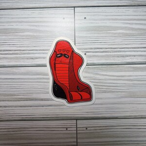 May include: A red racing seat sticker with black accents and three heart shapes at the top. The sticker has a white border and is placed on a light gray wooden surface.
