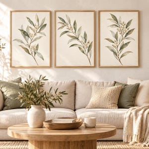 Puede incluir: Tres cuadros de pared enmarcados con ilustraciones en acuarela de ramas verdes con pequeñas flores blancas sobre un fondo crema, exhibidos en marcos de madera clara sobre un sofá beige con cojines decorativos verdes y beige.