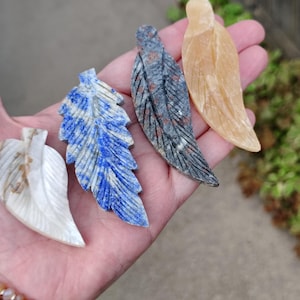 May include: Four carved stone leaf sculptures in various colors are displayed in a hand. The leaves are white, blue, gray, and yellow. The leaves have detailed veining and are smooth to the touch. The sculptures are a unique home decor item.