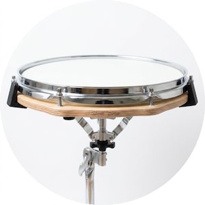 Könnte beinhalten: Ein Übungs-Drum-Pad mit einer weißen, kreisförmigen Oberfläche und einem metallischen Chromrand, montiert auf einer hellen Holzbasis mit dem Schriftzug Silent Drum. Die Trommel wird von einem verstellbaren silbernen Metallständer gehalten.