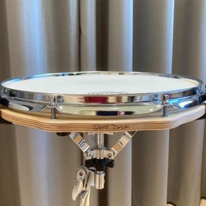 Op de afbeelding: Een oefenpad met een wit Remo drumvel, een verchroomde metalen rand en een lichtgekleurde multiplex basis. De woorden Silent Drum staan gedrukt op de zijkant van het houten frame. De trommel is gemonteerd op een zilverkleurige metalen standaard.