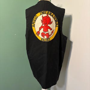 Peut inclure: Un gilet noir sans manches avec un écusson circulaire au dos. L'écusson représente un diable rouge tenant une fourche, entouré d'une bordure jaune avec le texte noir Apollo International Radio imprimé tout autour du bord.