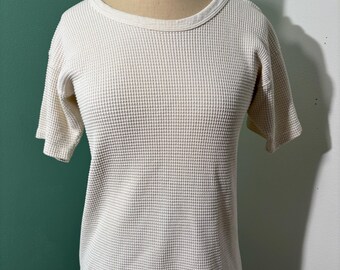 May include: Cream-coloured waffle-knit short-sleeved top on a mannequin. The shirt features a round neckline and a textured, grid-like pattern. The fabric appears to be a lightweight cotton or blend, suitable for casual wear. The hemline is slightly curved.