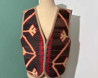 May include: A patterned waistcoat in brown and beige, with a V-neck. The design includes geometric shapes in red, beige, and dark brown. The waistcoat is displayed on a mannequin.