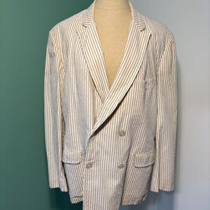 May include: Cream-colored blazer with vertical black pinstripes. The double-breasted jacket features two rows of buttons, notched lapels, and two front pockets. The blazer is displayed on a mannequin.