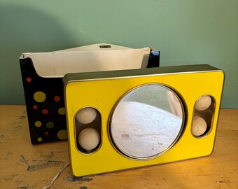 May include: A vintage yellow and gold vanity mirror with a round mirror and two sets of two white spheres. A black box with colorful polka dots sits behind the mirror. The mirror has a power cord.