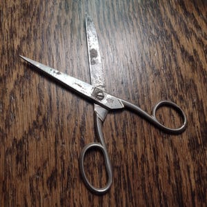 May include: Vintage metal scissors with a silver finish, resting on a wooden surface. The scissors have a classic design with loop handles and a screw at the pivot point. The blades show signs of age and use.