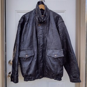 May include: Black leather jacket with a high collar, two front pockets, and a ribbed hem. The jacket is hanging on a wooden hanger against a white door. The jacket has a textured appearance.