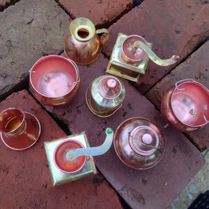May include: An assortment of small, copper-colored metal miniatures, including a coffee grinder, a small pitcher, a cup and saucer, and various bowls and containers. The items are arranged on a red brick surface.