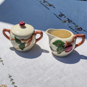 May include: Cream-colored ceramic sugar bowl with lid and creamer set. The set features a branch and fruit design with brown, green, and red accents. The handles are brown and the lid has a red knob. The creamer has a spout.