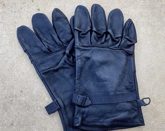 May include: A pair of navy blue leather gloves. The gloves have a textured appearance and feature a strap with a buckle closure around the wrist. The gloves are designed for protection and warmth.