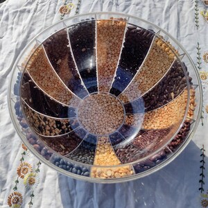 May include: A clear, circular container divided into pie-shaped sections, each filled with a different type of dried beans and lentils. The beans range in color from light tan to dark brown and black. The container sits on a white cloth.