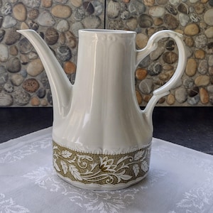 May include: A white ceramic coffee pot with a long spout and a curved handle. The base features a decorative band with a gold and olive green floral pattern. The pot is placed on a white patterned tablecloth.