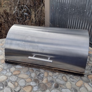 May include: A stainless steel bread box with a curved top and a rectangular base. The box has a silver handle and a dark brown wooden base. The bread box is designed to store bread and baked goods.