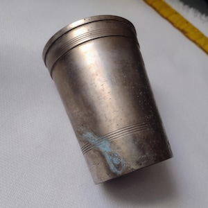 May include: A silver-colored metal tumbler with horizontal etched lines near the top and bottom. The cylindrical cup has a slightly tarnished appearance with some blue discoloration. The word "PRO" is partially visible on the side.