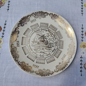 May include: A vintage white plate with gold trim and a calendar design for the year 1953. The plate features a windmill scene in the center and calendar boxes around the edge, with the months of the year.
