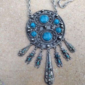 May include: Silver-toned statement necklace with turquoise-colored stones. The pendant features an oval design with multiple turquoise stones and dangling charms. The necklace has a chain and is displayed on a neutral surface.