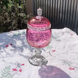 May include: A pink glass candy dish with a clear glass pedestal base and lid. The lid has a decorative finial and a patterned band around the rim. The dish is sitting on a white tablecloth with floral embroidery.