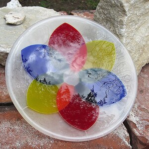 Pu&ograve; includere: Una ciotola di vetro trasparente con un design a forma di fiore. La ciotola presenta sei sezioni a forma di petalo in rosso, giallo e blu, ognuna delle quali riflette una scena di cielo e alberi. La ciotola &egrave; su una superficie di mattoni.