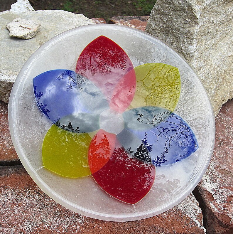 Pu&ograve; includere: Una ciotola di vetro trasparente con un design a forma di fiore. La ciotola presenta sei sezioni a forma di petalo in rosso, giallo e blu, ognuna delle quali riflette una scena di cielo e alberi. La ciotola &egrave; su una superficie di mattoni.