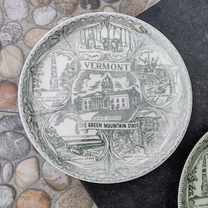 May include: A white ceramic plate with a green Vermont-themed design. The plate features illustrations of the State Capitol, a covered bridge, and other Vermont landmarks. The text "VERMONT" and "THE GREEN MOUNTAIN STATE" are also visible.